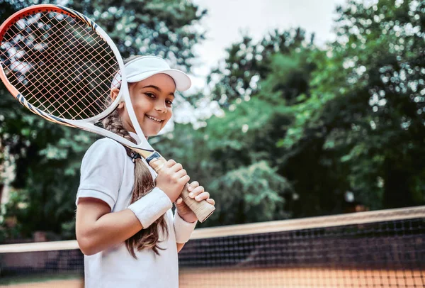 Tennis training for young kid outdoors. Portrait of happy sporty little girl on tennis court. Caucasian child in white tennis sportswear on training.