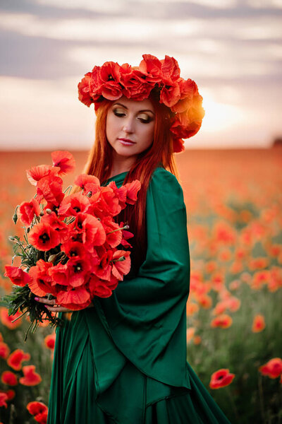 A young smiling red-haired woman in a wreath of poppies stands in a blooming poppy field. Medium shot.