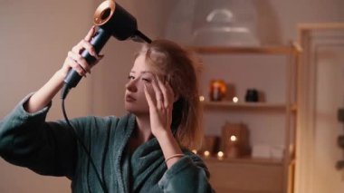 Bornoz giymiş bir kadın banyoda saçını saç kurutma makinesiyle kurutuyor..