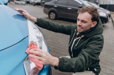 Gülümseyen adam açık hava otoparkında mikrofiber bir bezle mavi bir arabanın farını temizleyip parlatıyor..