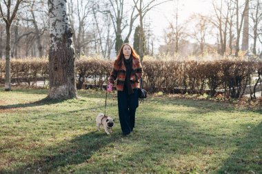 Genç bir kadın, sonbahar günü şehir parkında tasmalı küçük bir köpek gezdiriyor, açık hava aktivitelerinin ve evcil hayvan arkadaşlığının keyfini çıkarıyor.
