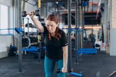 Odaklanmış genç bir kadın modern bir spor salonunda tek kollu bir halter egzersizi yapıyor. Antrenman gücü, denge ve tüm vücudunu kontrol ediyor..