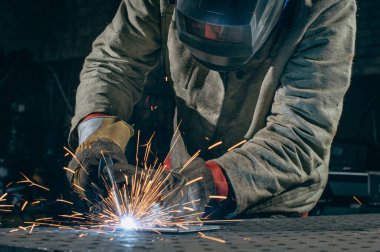 Profesyonel bir metal işçisi endüstriyel bir atölyede çelik kaynak yapar. Kaynak işleminden çıkan parlak kıvılcımlar gibi koruyucu giysiler giyer..