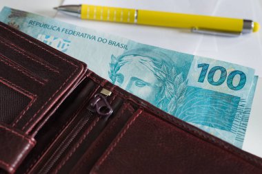 Brazilian banknotes, one hundred Reais sticking out of the wallet, a piece of paper with a pen next to it