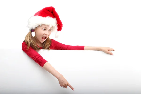 惊讶的小女孩，在圣诞老人的帽子偷看的显示一些孤立的白色背景上的白板上的肖像. — 图库照片