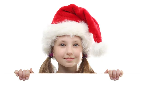 Портрет улыбающейся маленькой девочки в красной шляпе Санта-Клауса, смотрящей на белую доску и смотрящей на камеру на белом фоне . — стоковое фото
