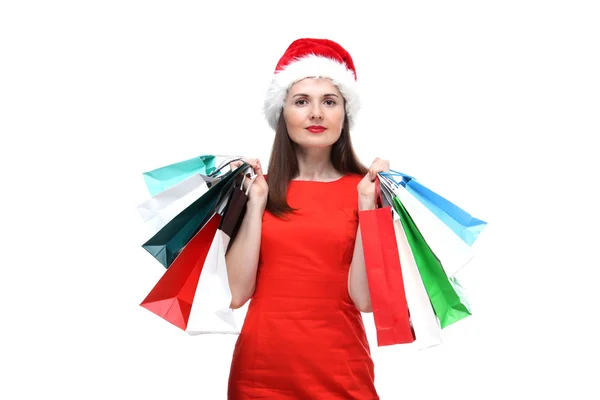 Портрет молодой взрослой красивой улыбающейся женщины в красном платье и шляпе Санта-Клауса, держащей цветные пакеты покупок изолированы на белом фоне — стоковое фото