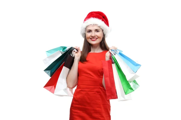 Портрет молодих дорослих красиві усміхається жінка носіння на червоній сукні і Санта-Клауса капелюх проведення кольорові сумками ізольовані на білому тлі — стокове фото