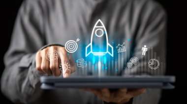 Parlak bir roket ikonu olan dijital tablet kullanan bir işadamı, modern dijital çağda başarı için başlangıç yeniliğini, iş açılışını, finansal büyümeyi ve yaratıcı teknoloji gelişimini sembolize ediyor..