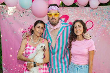 Pembe süslemeleri, balonları ve renkli kıyafetleri olan küçük beyaz bir köpeği olan bir partide fotoğraf için poz veren aile.