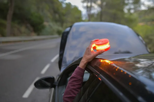 Hand placing a V16 emergency safety warning beacon on a broken down car with its hood open, ensuring roadside visibility and safety during a breakdown