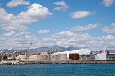 Fabrika kompleksi ve dağlık gökyüzü altında sanayi limanı manzarası.