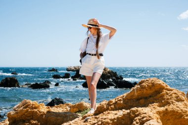 Yaz kıyafeti ve şapkasıyla kayalık sahili keşfeden bir kadın..
