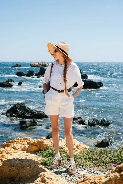 Beyaz elbiseli ve hasır şapkalı kadın kayalık kıyı şeridinde manzaranın tadını çıkarıyor..