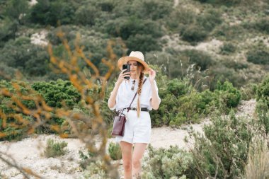 Beyaz şapkalı ve beyaz elbiseli akıllı telefonlu bir kadın doğayı keşfediyor..