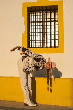 Şık batı kıyafetli kadın gün batımında bükülüyor sarı ve beyaz bir duvarın altında..