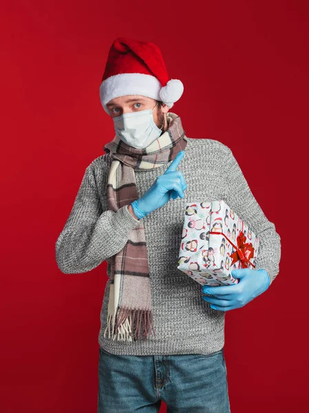 A young man in a medical mask and gloves, wearing a Santa hat and scarf holds a gift box and points to the upper-right corner on a red background