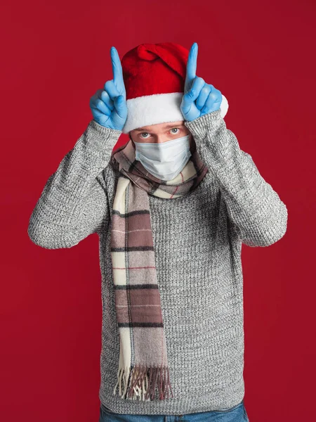 A young man in a medical mask and gloves, wearing a Santa hat and a scarf, acting out a bull