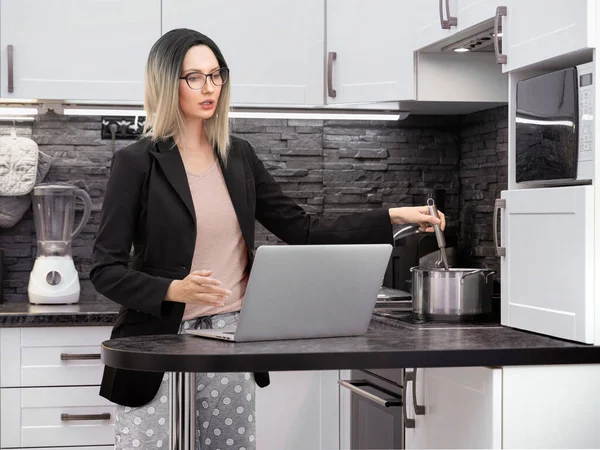 Blonde girl in glasses, black formal jacket and pajama pants conducts business talks on the Internet via a laptop in the kitchen and stirring in a pot on the stove, works remotely at home