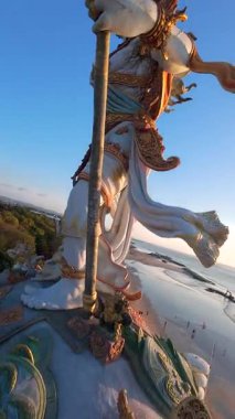 Endonezya, Bali 'de gün batımında görkemli okyanus manzaralı heykelin etrafında uçan FPV dronu. Adaların altın ışığı, dalgaları ve kültürel güzelliği..