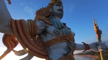 Endonezya, Bali 'de gün batımında görkemli okyanus manzaralı heykelin etrafında uçan FPV dronu. Adaların altın ışığı, dalgaları ve kültürel güzelliği..