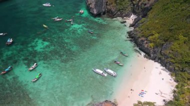 Turkuaz su, kayalıklar ve yemyeşil yeşillik ile çevrili beyaz kumlu sahilde demirli teknelerin olduğu tropik körfezin sinematik hava manzarası. Seyahat, tatil ve cennet temalı projeler için mükemmel..