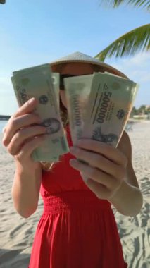 Vietnam 'da palmiye ağaçlarının altındaki tropik plajda Vietnamlı dong banknotlarını sayan kırmızı elbiseli bir kadın. Asya 'da zenginlik ve özgürlük hakkında seyahat, finans ve yaşam tarzı kavramları için mükemmel..