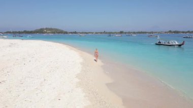 Endonezya 'daki Gili Adaları' ndaki turkuaz okyanus kıyısında kumlu tropikal sahilde yürüyen bir kadının hava görüntüsü. Berrak su, egzotik sahil ve tatil atmosferli yaz seyahati sahnesi.