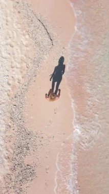 Kumlu tropik plajda kristal berrak deniz ve gölgesi kıyı şeridi boyunca yürüyen kadının yukarıdan aşağı görüntüsü. Yaz, tatil ve kıyı yaşam tarzının minimalist ve sanatsal sahnesi.