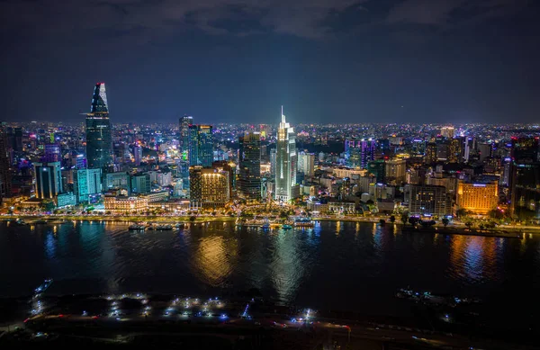 Ho Chi Minh City, Vietnam 'ın gece hava görüntüsü, aydınlık gökdelenler, nehir kenarındaki ışıklar, ve canlı şehir silueti suya yansıyor..