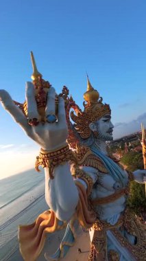 Endonezya, Bali 'de gün batımında görkemli okyanus manzaralı heykelin etrafında uçan FPV dronu. Adaların altın ışığı, dalgaları ve kültürel güzelliği..