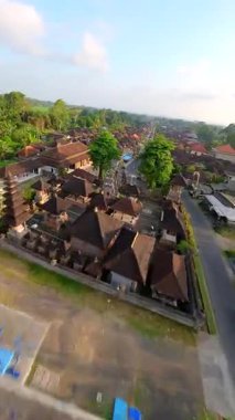 Endonezya 'nın Bali kentindeki geleneksel köy üzerinde FPV insansız hava aracı uçuşu, yerel tapınağın üzerinde başlıyor ve bereketli tropikal yeşil evler ve köy evleriyle çevrili süslü cadde boyunca devam ediyor..