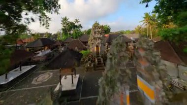 Endonezya 'nın Bali kentindeki geleneksel köy üzerinde FPV insansız hava aracı uçuşu, yerel tapınağın üzerinde başlıyor ve bereketli tropikal yeşil evler ve köy evleriyle çevrili süslü cadde boyunca devam ediyor..