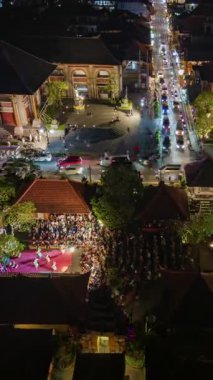 Ubud, Bali 'nin merkezinde yoğun sokak trafiği, aydınlık binalar ve tropikal bahçeler ve canlı gece ışıkları ile çevrili Ubud Sarayı' nın yakınında görünür bir açık hava sahnesi var..