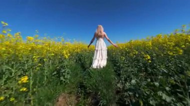 Güneşli yaz gününde sarı kolza tohumu tarlasında yürüyen bir kadının sinematik FPV görüntüsü. Doğa, özgürlük ve yavaş yaşam temaları için huzurlu kırsal yaşam tarzı sahneleri.