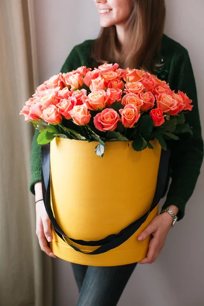 Nice young blonde girl. smelling flowers holding peach roses bouquet in hat box against the plastered wall, wearing jeans knit sweater.