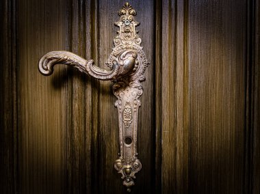 Zengince detaylandırılmış eski bir kapı kolu ve altın kaplamalı anahtar deliği plakası, koyu, desenli ahşap bir kapıya dayanıyor. Tarihi, lüksü ve güvenliği simgeliyor.