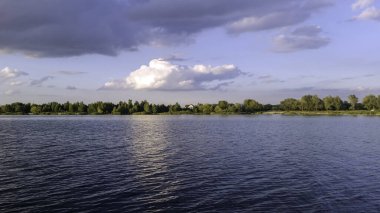 Ufukta yemyeşil bir orman hattı tarafından çerçevelenmiş pofuduk beyaz ve gri bulutlarla güzel bir gökyüzünün altında sakin bir gölün panoramik manzarası. Barışçıl doğal çevre.