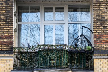 Dekoratif işlenmiş demir parmaklıklar, büyük kuş kafesi ve modern beyaz çerçeveli pencerelerde çıplak ağaçların yansımaları olan eski apartman dairesinin ön manzarası.