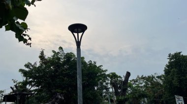 Bu sakin park sahnesiyle doğaya kaçmak. Huzurlu bir gökyüzünün altındaki yemyeşil ağaçların arasında parıldayan bir ışık direği duruyor. Huzur ve rahatlama için mükemmel bir yol..