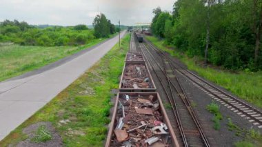 Yeşil yol kenarındaki paralel raylar boyunca uzanan hurda metal parçalı uzun açık vagonlar. Tren seti asfalt yolu ve kargo başlıklı küçük direklerden uzaktaki tesise doğru ilerliyor.
