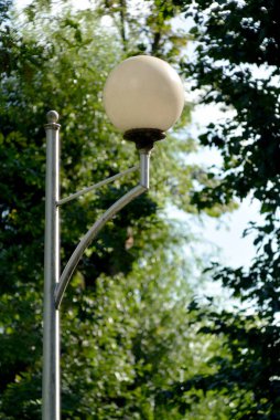 Yuvarlak mat küreli klasik bir sokak lambası yoğun yeşil yaprakların arasında duruyor. Doğal olarak sakin ve huzurlu park atmosferine karışıyor..