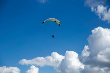 Bir paraglider gökyüzünde süzülüyor, beyaz bulutların arasında, derin mavi bir zemine karşı uçma özgürlüğü ve sakinliğine daldırılmış..