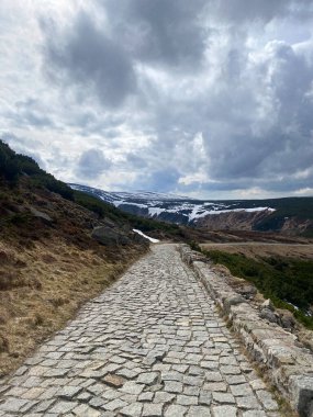 Sniezka 'dan inen taş bir patika Karkonosze yamaçları arasında esen rüzgarlar dramatik bulutlu bir gökyüzünün altındaki karlı tepeleri ortaya çıkarıyor. Sahne sakin ve görkemli..