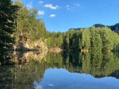 Sakin bir göl, yeşil ormanı, beyaz bulutları ve kayalık kayalıkları yansıtır ve mükemmel bir simetrik ve pitoresk doğal manzara yaratır..
