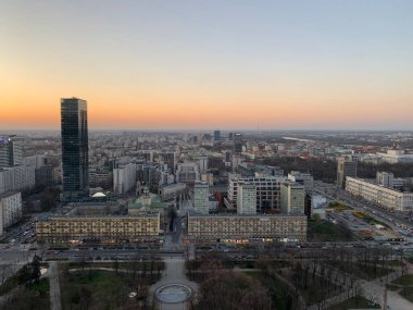Evening Warsaw from above: warm sunset tones, modern buildings, dense urban landscape, and a calm horizon stretching beyond the city center.