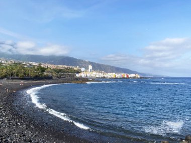 Siyah volkanik plajlı Tenerife kıyısı, Atlantik Okyanusu dalgaları ve kıyı kasabası manzarası. Dağlar, bulutlar ve mavi su. Kanarya Adaları 'nın gezisi ve doğası.
