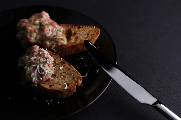 Pyszne tosty z bagietką do chleba z rybami, zielonymi, serem i sosem na czarnej płytce w pobliżu noża, ciemne tło — Zdjęcie stockowe
