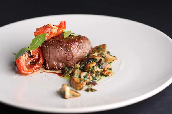 Жареное мясо с помидорами и грибами на белой тарелке, темный фон в ресторане — стоковое фото