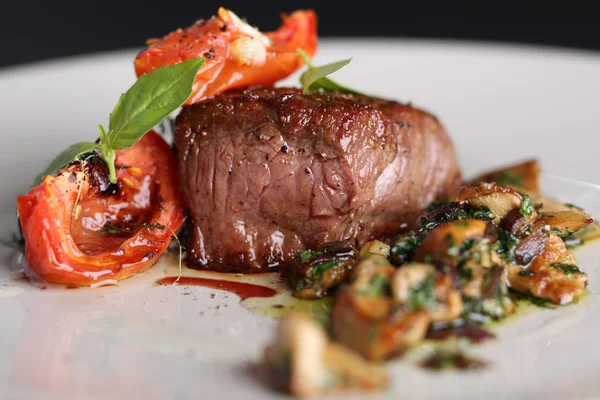 Viande rôtie avec des tomates et des champignons sur une assiette blanche, fond sombre dans le restaurant. Gros plan . — Photo